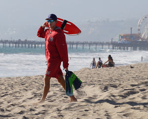 Male lifeguard on the beach in a red jacket and swim trunks wearing blue nose sunscreen is holding a rescue buoy and getting ready to go in the water.